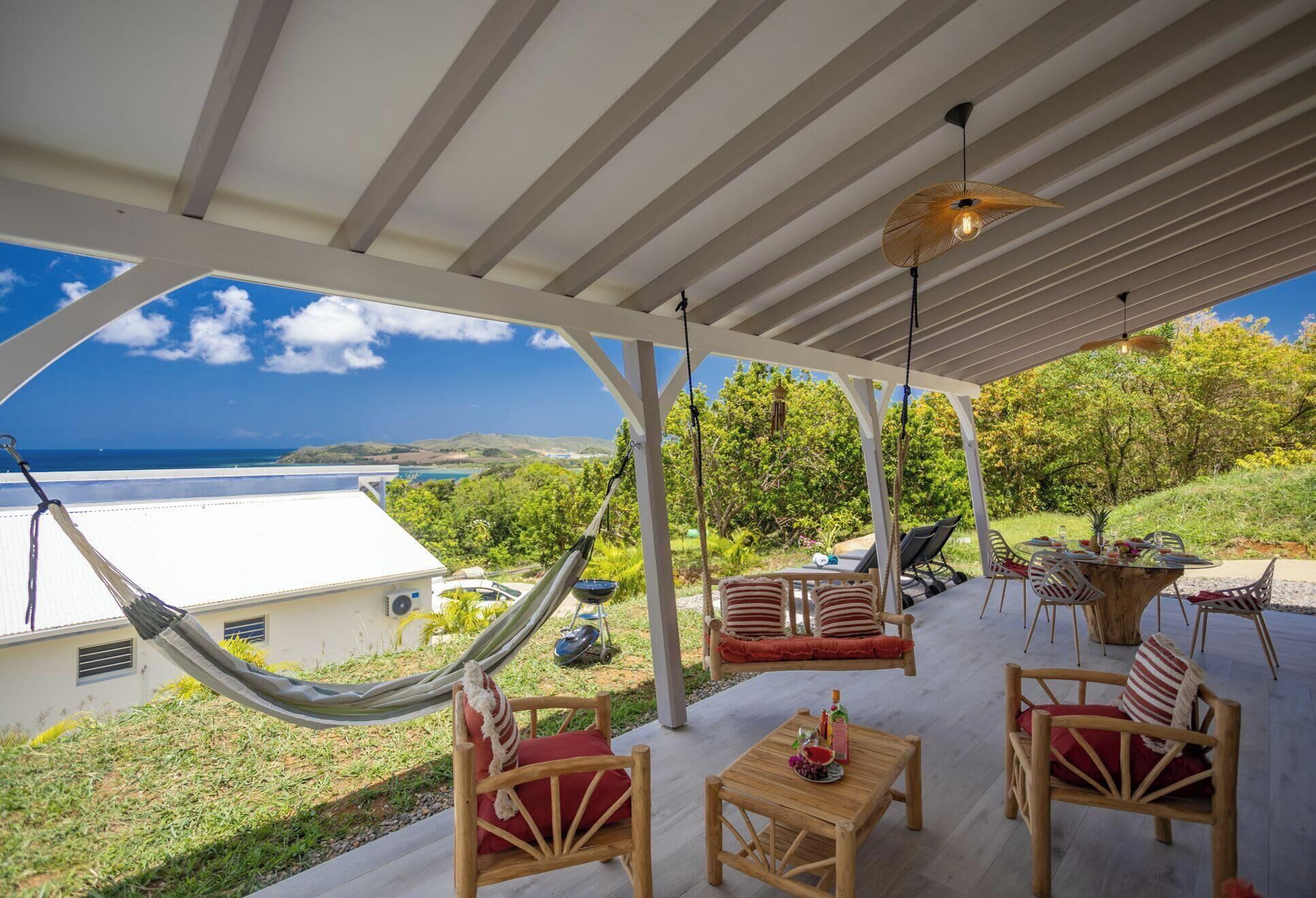 Un área de estar techada al aire libre con una hamaca, sillas de madera y una mesa, con vistas a un paisaje pintoresco y al mar y las colinas.