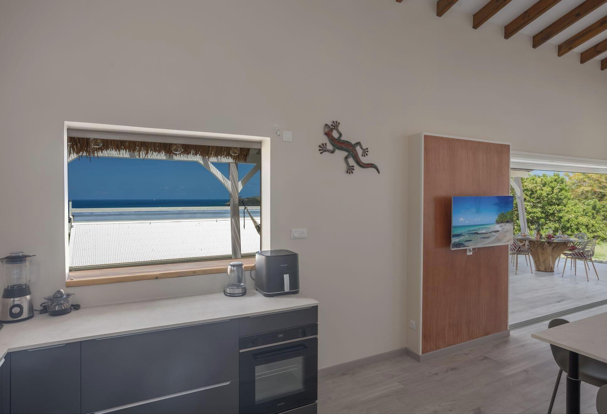 Une cuisine moderne dotée d’une grande fenêtre donnant sur une plage, d’un meuble en bois orné d’un tableau, et d’un coin repas avec vue sur la verdure.