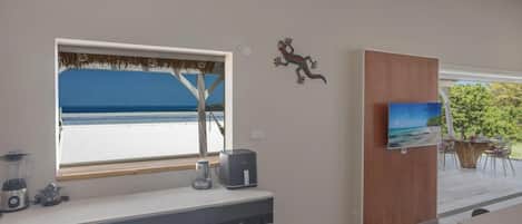A modern kitchen with a large window overlooking a beach, a wooden cabinet with a painting, and a dining area with a view of greenery.