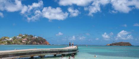 Une plage à l’eau turquoise et limpide, une jetée en bois et, au loin, des bâtiments situés sur une colline.