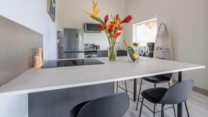 A modern kitchen with a central island, a dining area with chairs, and a surfboard shelf. - La Villa de Solange - Azur (La Trinité)