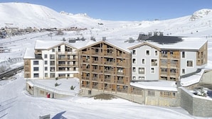 Un hotel de varios pisos con fachada de madera, situado en una zona montañosa nevada con pistas de esquí al fondo.