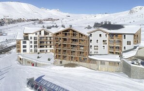 A multi-story hotel with a wooden facade, located in a snowy mountainous area with ski slopes in the background. - T2 au pied des pistes | Piscine sur place | Garage (Alpe d'Huez)