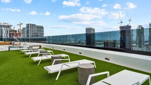 Terraza en la azotea con sillones blancos y barandilla de cristal, con vistas a un paisaje urbano con edificios modernos y grúas de construcción.