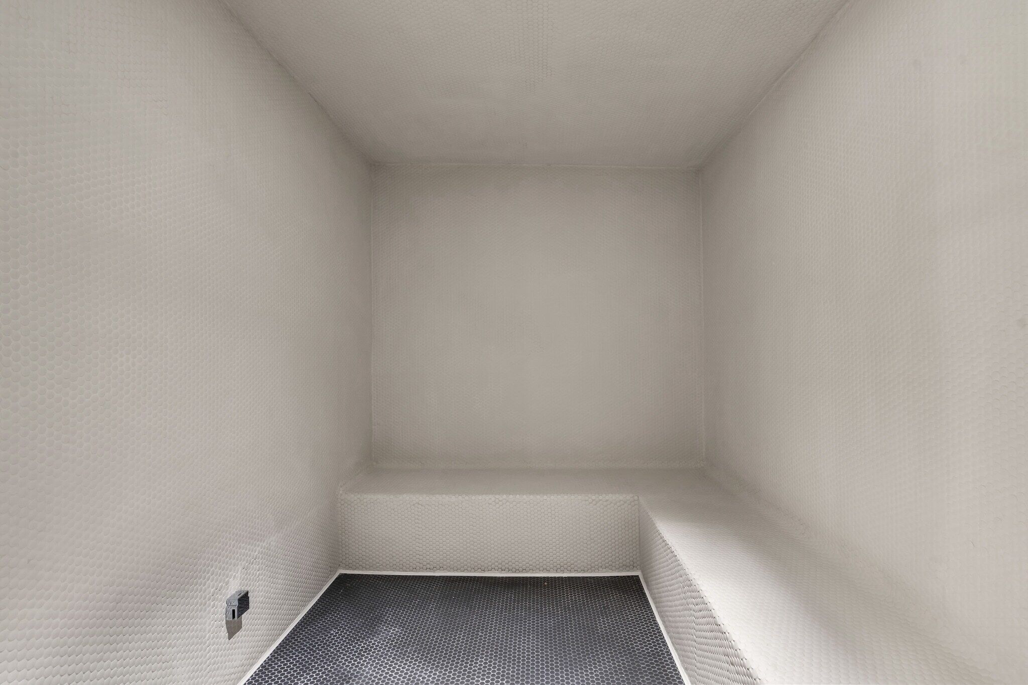 Une pièce avec un mur blanc à la texture particulière, un sol à motifs noir et blanc et une petite grille de ventilation.