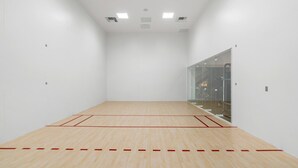Una cancha de squash con suelo de madera, paredes de vidrio e iluminación en el techo.