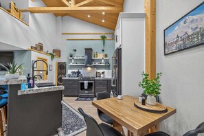 A modern kitchen with dark cabinetry, a central island, and a dining area with a wooden table and chairs. - Hot Tub Barndominium Near Sunriver & Deschutes (Bend)