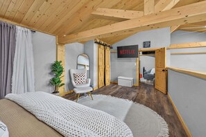 A room with a bed, a white armchair, a TV, and a wooden ceiling. - Hot Tub Barndominium Near Sunriver & Deschutes (Bend)