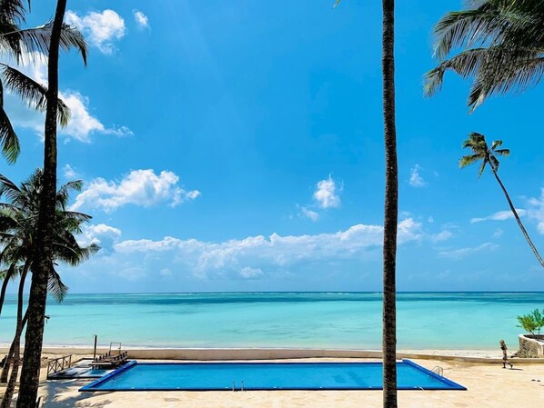 Céu azul límpido, piscina e praia com palmeiras.