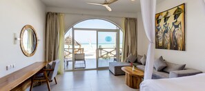 Quarto com cama, escrivaninha de madeira, sofá e um quadro. Tem vista para a praia através de uma porta de vidro.