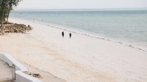 Uma praia com areia branca, algumas pessoas caminhando e um mar calmo ao fundo.