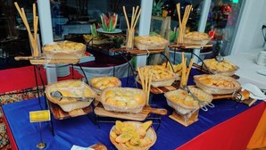 Mesa de buffet com diversos tipos de pão e doces, alguns cestos com palitos de pão e uma placa indicando os alimentos disponíveis.