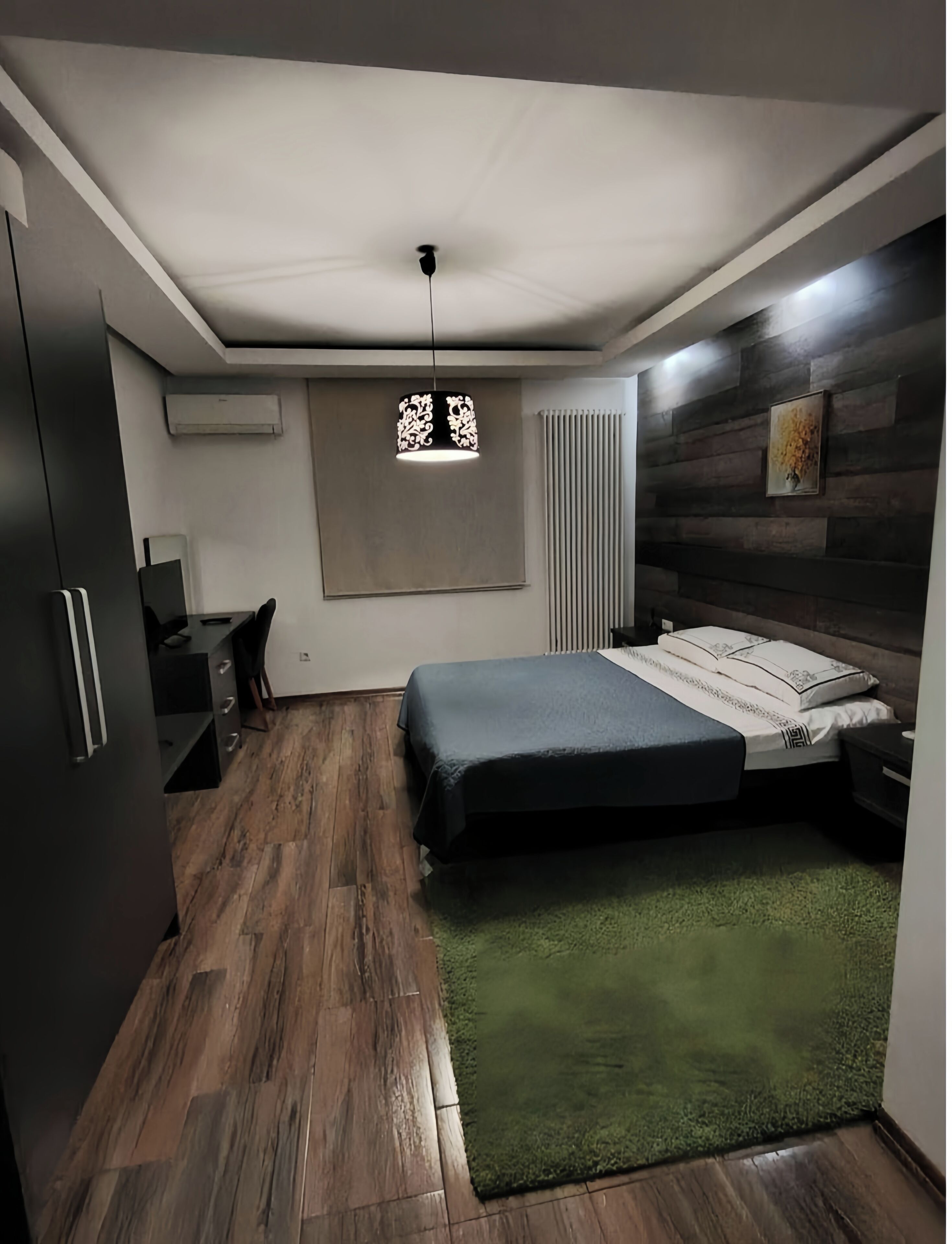A bedroom with a bed, a desk, a chair, a wardrobe, a radiator, and a hanging light fixture.