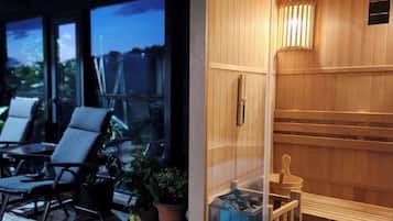 Un sauna avec un intérieur en bois et une porte en verre, situé à côté d’un espace piscine comprenant des chaises longues et des plantes en pot.