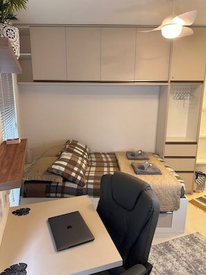 Ein gemütliches Schlafzimmer mit einem Bett, einem Schreibtisch mit Laptop, einem Stuhl, einem Deckenventilator und einem Kleiderschrank.
