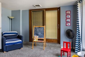Ein Zimmer mit blauem Sessel, einem kleinen hölzernen Staffelei mit einer Kreidetafel, einem roten Stuhl, einem Ventilator und einem Fenster mit Jalousien.