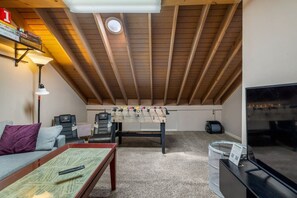 Dormitorio tipo loft con techo de madera, mesa de fútbol, sofá, televisor y pequeña mesa con un control remoto.