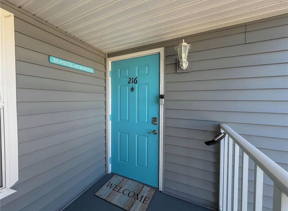 Une porte turquoise portant le numéro 216, un paillasson et une enseigne « Souvenirs de bord de mer » sur la façade d’un immeuble.