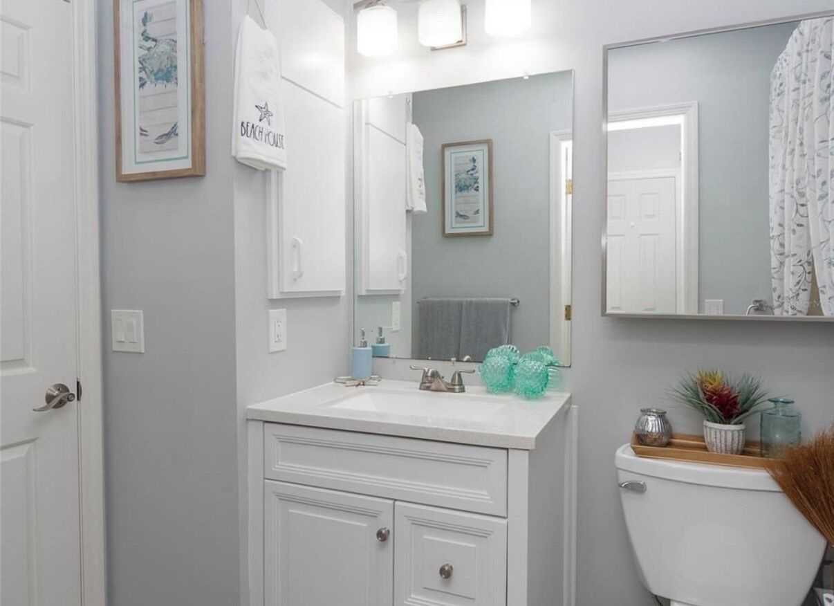 A bathroom with a white vanity, a mirror, and a toilet. There are framed pictures on the wall and a towel hanging on the door.