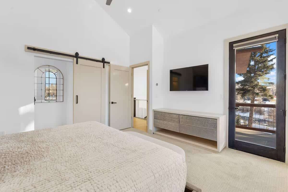 A bedroom with a bed, a sliding door, a TV mounted on the wall, and a view through a door to the outside.