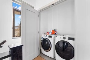 A laundry room with two washing machines, a small table, and a window with a view of the outside. - NEW! The Vista On The Blue - Downtown Riverfront (Silverthorne)