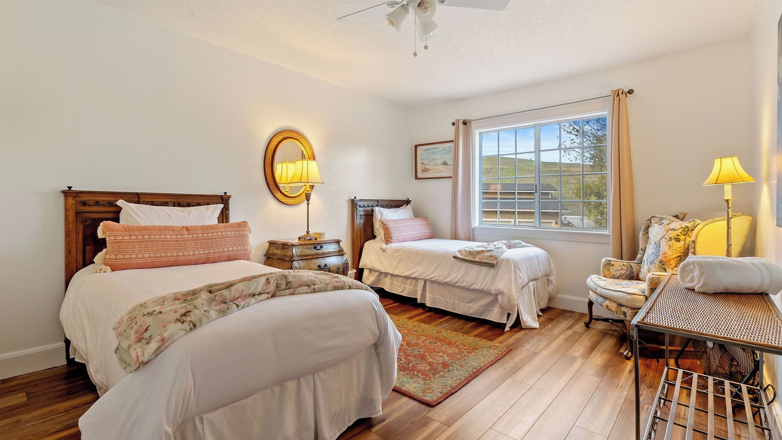 A bedroom with two beds, a large window, a ceiling fan, and a chair with a lamp.