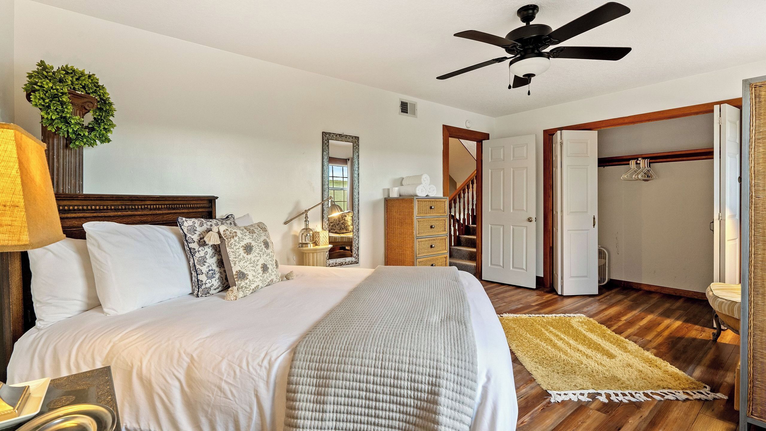 Quarto com cama, ventilador de teto, mesa de cabeceira, cômoda, espelho e porta para outro espaço.