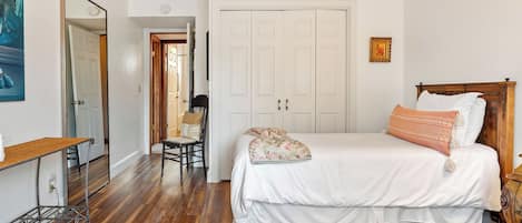 A bedroom with a wooden floor, a bed with white bedding, a chair, a desk, and a wardrobe.