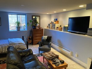 A living room with a bed, a leather sofa, a coffee table with remotes, a TV, a guitar, and a dresser with shoes. - Garden level studio apartment downtown Boulder (Boulder)