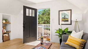 A modern living room with a dark door, a framed poster, a floor lamp, a sofa with cushions, a coffee table with a wine bottle and glasses, and a bookshelf with various items. - The Nook on Hilltop by Avantstay Skylit Loft + Private Entry + Induction Kitchen Walk to Downtown Paso Robles Wine Country (Paso Robles)