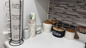 Een keukenblad met een koffiemokkenhouder, gestapelde koffiekopjes en gelabelde bakjes voor koffiefilters, room en suiker.