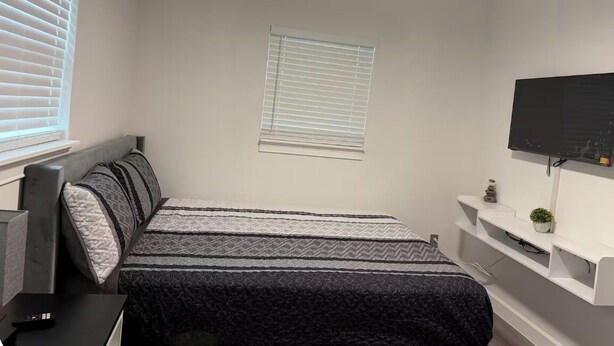 Een slaapkamer met een bed, een aan de muur bevestigde televisie, een plafondventilator en een raam met zonwering.
