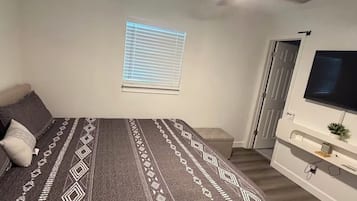 Um quarto com uma cama, um ventilador de teto, uma janela com persianas, uma porta e uma televisão fixada na parede.