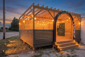 Una estructura de madera con techo a listones y luces colgantes, una pequeña entrada con escalones y un arco decorativo.