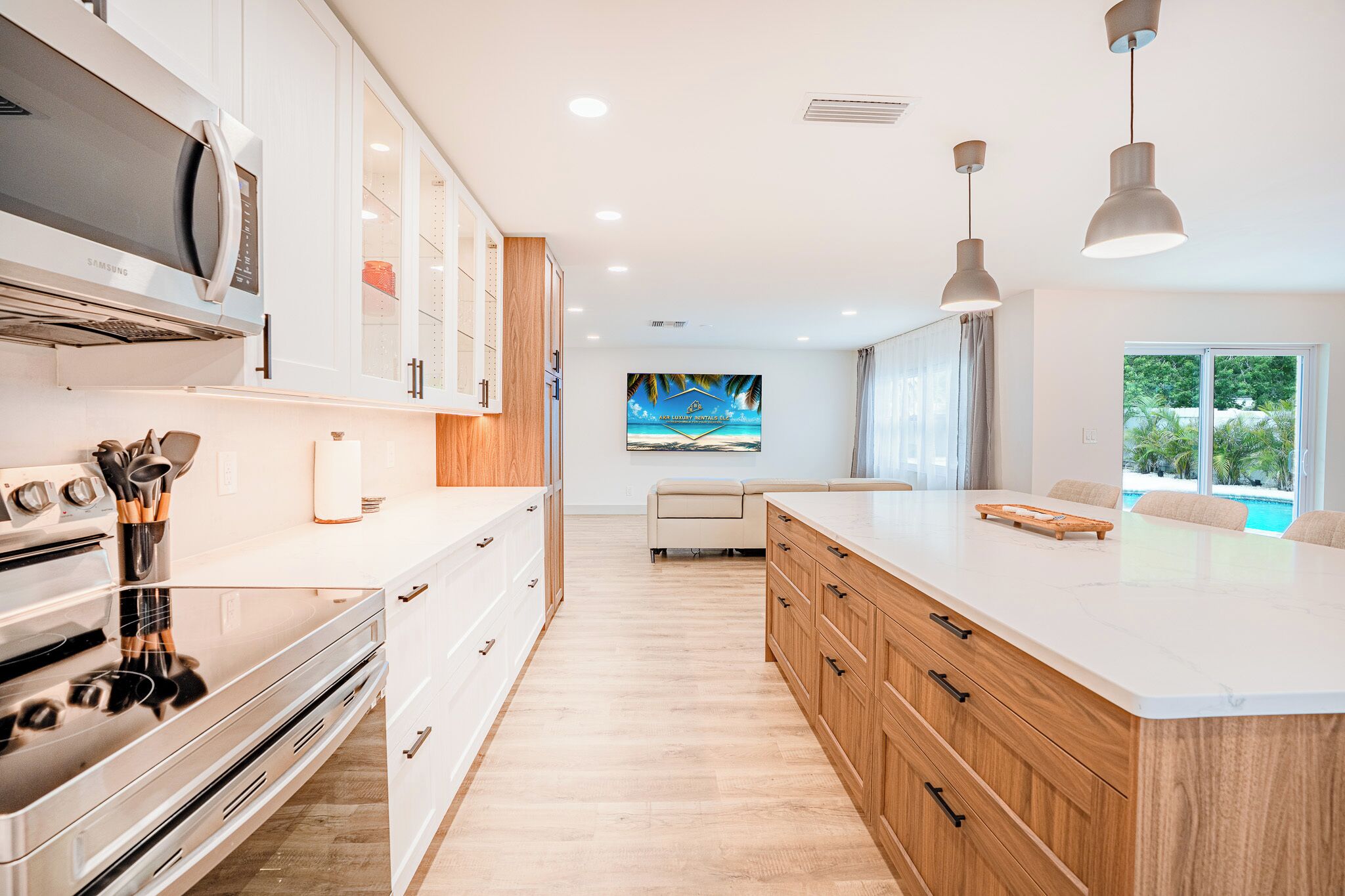 A modern kitchen with a central island, stainless steel appliances, wooden cabinets, and a large window with a view of a pool.