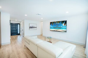 Un salon moderne avec un canapé blanc, une petite table en verre et un téléviseur fixé au mur qui diffuse une image d’une plage tropicale.