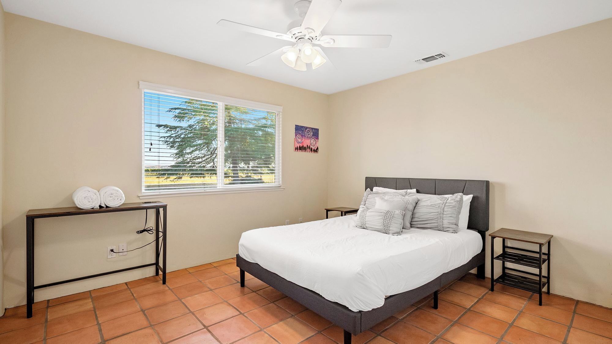 A bedroom with a bed, a desk, a chair, a ceiling fan, and a window with a view of trees.