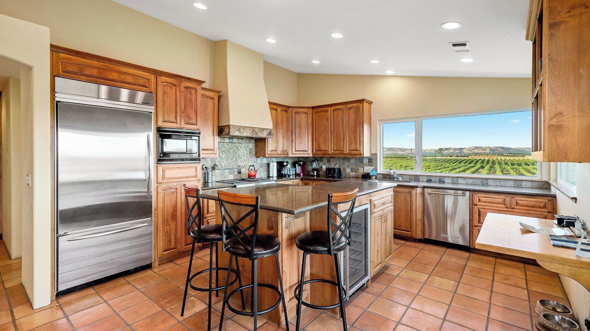 A kitchen with a large island, stainless steel appliances, wooden cabinets, and a view of a vineyard.