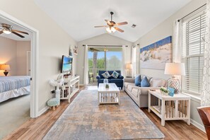 A spacious living room with a ceiling fan, a large window with blinds, a sofa, a coffee table, and a TV mounted on the wall. - River Oaks 49I Myrtle Beach | Great Fall Price! (Myrtle Beach)
