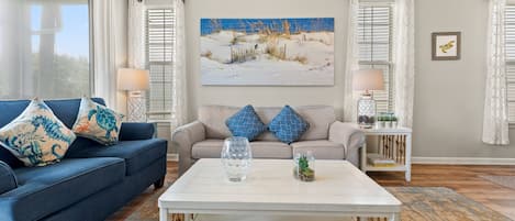 Uma sala de estar com um sofá azul, duas almofadas estampadas, uma mesa de centro de vidro, uma pintura de uma cena de praia e uma pequena planta sobre uma mesinha lateral.