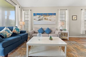A living room with a blue sofa, two patterned pillows, a glass coffee table, a painting of a beach scene, and a small plant on a side table. - River Oaks 49I Myrtle Beach (Myrtle Beach)