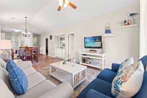 A spacious living room with a white sofa, blue sofa, and a coffee table. There is a dining area with a wooden table and chairs. A TV is mounted on the wall, and there are decorative items on shelves. - River Oaks 49I Myrtle Beach (Myrtle Beach)