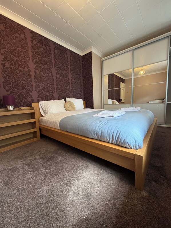 Une chambre à coucher avec un lit en bois, une table de chevet, un miroir et un papier peint à motifs.