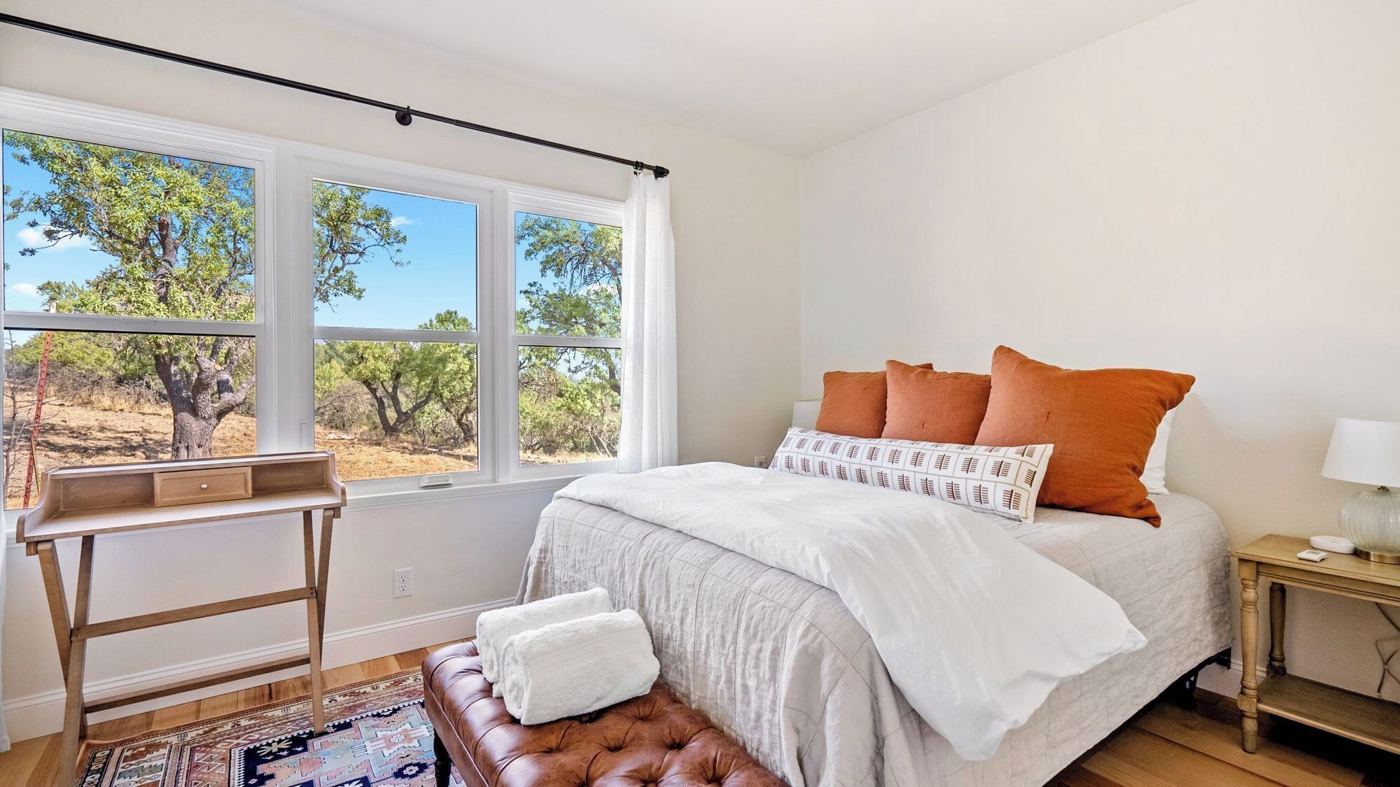 A bedroom with a large window, a bed with multiple pillows, a bedside table, and a bench.