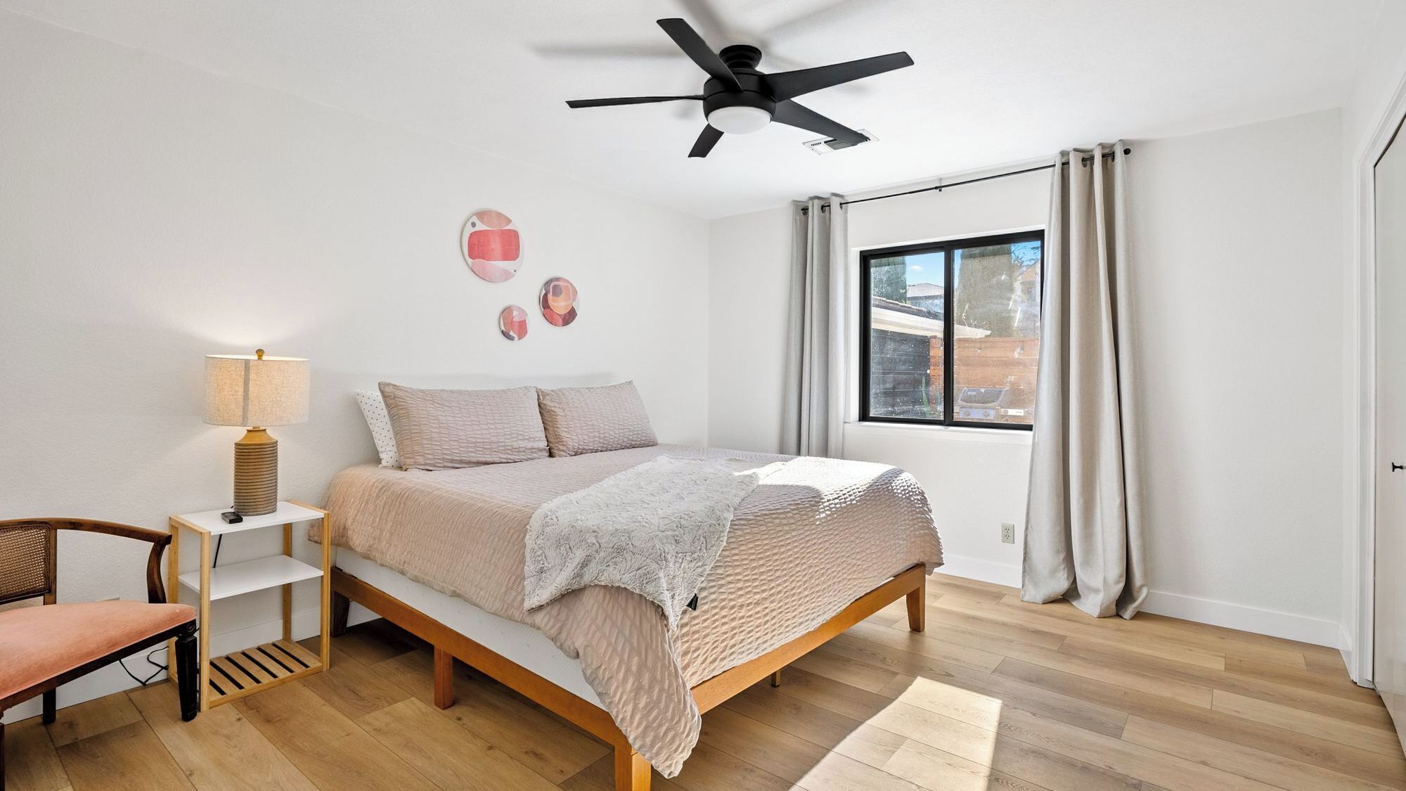 A bedroom with a bed, a chair, a bedside table, a lamp, a ceiling fan, and a window with curtains.