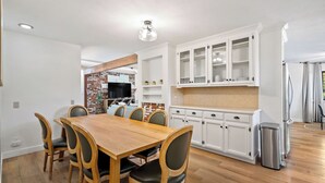A dining area with a wooden table and chairs, white cabinetry with glass doors, and a kitchen area with a brick accent wall. - Prickly Pear Paso by Avantstay Fire Pit + BBQ Patio Walkable Downtown Paso (Paso Robles)