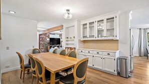 A dining area with a wooden table and chairs, white cabinetry with glass doors, and a kitchen area with a brick accent wall. - Prickly Pear Paso by Avantstay Fire Pit + BBQ Patio Walkable Downtown Paso (Paso Robles)
