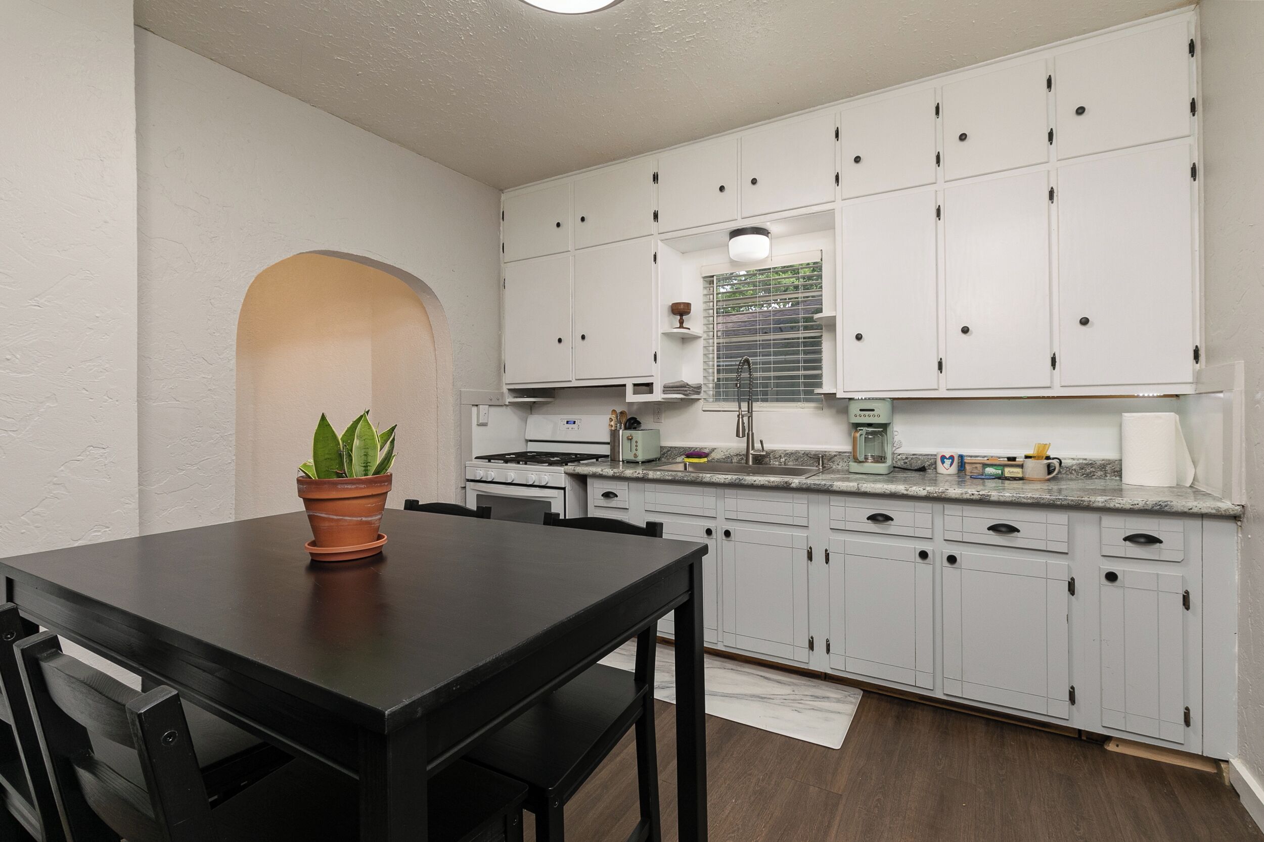 Una cocina con armarios blancos, un comedor con mesa negra y una planta en maceta.