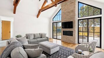 A spacious living room with a high ceiling, a large sofa set, a fireplace, and large windows offering a view of the outdoors.
