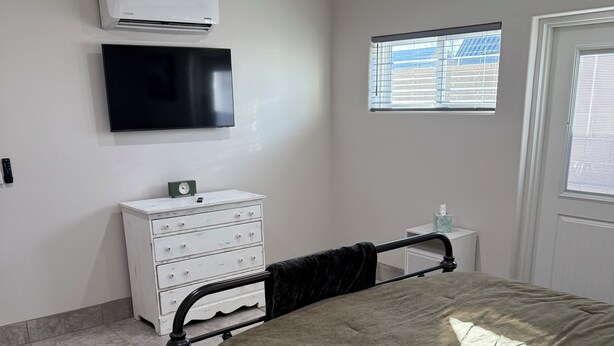 A bedroom with a bed, a white dresser, a television, an air conditioner, a ceiling fan, and a window with blinds.